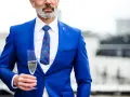 Elegancki mężczyzna w niebieskim garniturze i wzorzystym krawacie, trzymający kieliszek szampana. Idealny strój koktajlowy na specjalne okazje.