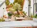 Mann hält Topf mit lila Blumen, um seine Dachterrasse mit Pflanzen zu gestalten. Eine Frau sitzt am Tisch.