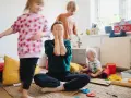 Chaos im Kinderzimmer. Eine Mutter versteckt ihr Gesicht in den Händen, während ihre Kinder spielen. Trotz des Trubels ist die liebe zu kindern spürbar.