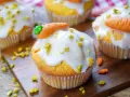 Muffins mit weißer Glasur, gehackten Pistazien und Marzipan-Karotten auf Holzbrett.