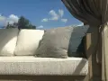 Wygodna sofa z poduszkami, osłonięta zasłonami, stoi na tle błękitnego nieba. Jej rama wykonana jest z pięknego, naturalnego **drewna tekowego**.
