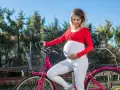 Kobieta w zaawansowanej ciąży, ubrana na biało i czerwono, z uśmiechem na twarzy, opiera się o różowy rower. Czy w ciąży można jeździć na rowerze? Tak, jeśli czujesz się dobrze i zachowasz ostrożność.