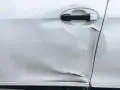 Widoczne wgniecenie i zarysowania na srebrnych drzwiach samochodu, świadczące o uderzeniu drzwiami na parkingu.