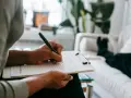 Osoba pisze na kartce, planując jak odmówić spotkania. W tle widać kanapę i rośliny.
