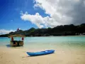 A blue kayak rests on a sandy beach in the Cook Islands, with a thatched-roof boat and lush green mountains in the background.