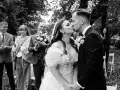 Para Młoda całuje się podczas ceremonii ślubnej. W tle widać gości, w tym pana młodego z bukietem. f6.