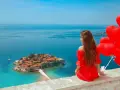 Marzenie o czarnogóra all inclusive samolotem? Kobieta w czerwonej sukni z sercami w ręku podziwia wyspę Sveti Stefan.
