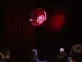 Na scenie, wśród czerwonych zasłon, unosi się kula z projekcją twarzy, przypominająca kurkę wodną. Widownia wpatruje się w to widowisko.