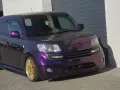 Fioletowy Daihatsu Materia z błyszczącymi złotymi felgami, zaparkowany przed garażem.