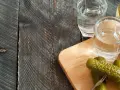 Zwei Gläser Wodka und eingelegte Gurken auf einem Holzbrett. Perfekt für alle, die Wodka selber machen wollen.