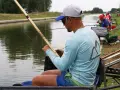 Wędkarz w czapce i okularach przeciwsłonecznych łowi ryby na tyczkę przy brzegu kanału.
