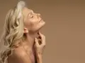 Kobieta z blond włosami i zamkniętymi oczami delikatnie dotyka szyi. Jej skóra w okresie menopauzy jest widoczna, podkreślając naturalne piękno.