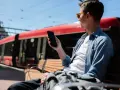 Mężczyzna w okularach siedzi na ławce i trzyma telefon. W tle czerwony tramwaj.