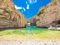 Spiaggia di ciottoli e acque cristalline tra le rocce dell'isola di Lissa, con persone che nuotano e barche.