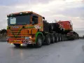 Pomarańczowy Scania z naczepą przewozi koparkę. Transport ponadgabarytowy wymaga specjalnych godzin jazdy.