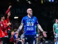 Trener SG Flensburg-Handewitt w niebieskiej koszulce z tatuażem na ramieniu, z otwartymi dłońmi, podczas meczu.