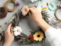 Hände gestalten einen Blumenkranz mit Heidekraut, Gerbera und Baumwolle. Das Binden eines Kranzes ist eine kreative Auszeit.