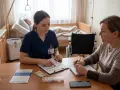 Pielęgniarka paliatywna omawia z pacjentką wyniki badań. W tle łóżko szpitalne i wózek inwalidzki.
