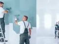 Malowanie ścian na miętowo. Dwóch fachowców na drabinie i stojącego maluje, trzeci pracuje przy stole. Sprawdź, ile za metr malowania ścian!