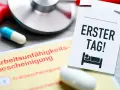 Arbeitsunfähigkeitsbescheinigung und eine Notiz "Erster Tag!" mit einem Bett-Symbol. Ein Stethoskop und Pillen deuten auf eine Krankschreibung hin, um gesund schreiben zu lassen.