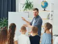 Nauczyciel tłumaczy coś dzieciom przy tablicy. To mogą być dni dyrektorskie, podczas których uczniowie poznają szkołę.