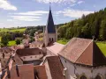 Vue aérienne d'un village pittoresque du haut Doubs, dominé par une église à la flèche élancée, entouré de collines verdoyantes et de forêts.