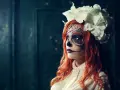 Kvinna med detaljerad ansiktsmålning för halloween, klädd i en vit blus och med en stor blomsterkrans.
