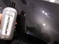Bottle of "Afterglow Metallic Infused Wax" held against a dark, metallic car finish, showcasing the product's ability to create multiple coats of wax for a deep shine.