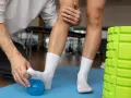 Fizjoterapeuta pomaga w rehabilitacji stawu skokowego, masując stopę pacjenta niebieską piłeczką. Obok stoi zielony wałek do masażu.