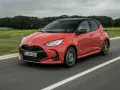 Auto ibrida rossa in movimento su strada, con sfondo sfocato di alberi e cielo nuvoloso. La scritta "Hybrid" è visibile sul fianco.