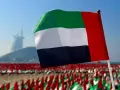 Flaga Zjednoczonych Emiratów Arabskich powiewa na tle drapaczy chmur w Dubaju. W tle widać morze małych flag.
