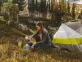 A woman enjoys a peaceful moment of camping alone, preparing a warm drink next to her tent amidst a serene forest landscape.