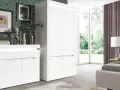 Nowoczesna szafa Irma w białym połysku, obok komody, na tle zielonej ściany z geometrycznym wzorem.