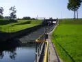 Zapora Mylof z kaskadami schodzącymi do wody. Po prawej stronie zielone, trawiaste zbocze z alejką i barierkami.