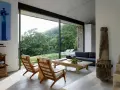 Salón moderno con sofás, sillones y mesa de centro rústica. Los suelos de piedra, con un estilo que evoca los años 70, contrastan con la vista exterior de un bosque frondoso.