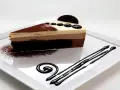 Una rebanada de tarta de tres chocolates, con capas de chocolate blanco, con leche y negro, decorada con cacao en polvo y un disco de chocolate.