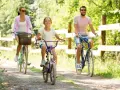 Rodzina cieszy się rodzinnym wypadem na **Mazury rowerem**. Radosna dziewczynka jedzie na fioletowym rowerze, a za nią rodzice.