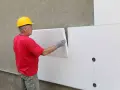 Operaio con casco giallo applica pannelli isolanti bianchi. Attenzione: il polistirolo è infiammabile.