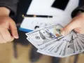 Dłoń trzyma plik banknotów 100-dolarowych, symbolizujących darowiznę od rodziców. Na stole leży długopis i dokument.