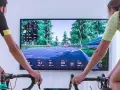 Two cyclists race virtually on a scenic road using indoor cycling apps. The screen displays their stats and rankings.