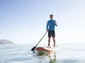 Mann steht auf SUP-Board, hält Paddel richtig. Ruhige See, bergige Küste im Hintergrund.