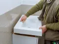 Installation d'un lavabo blanc. L'installateur vérifie la hauteur de l'arrivée d'eau du lavabo avant de le fixer.
