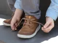 Kind lernt, seine braunen Schnürstiefel zu binden. Die Schnürsenkel sind hellbraun und gemustert.