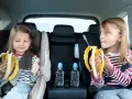 Dwie dziewczynki w fotelikach samochodowych jedzą banany. Woda i przekąski to podstawa, gdy zastanawiasz się, co zabrać na długą podróż samochodem z dzieckiem.