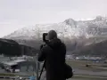 Mężczyzna w czapce i kurtce fotografuje krajobraz z ośnieżonymi górami. Widać przemysłową zabudowę i tory kolejowe. To jego sposób na uchwycenie **mark power**.