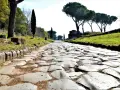 Ancient Roman cobblestone road lined with trees and ruins, leading towards the horizon.