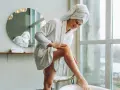 Frau in Bademantel und Handtuch auf dem Kopf pflegt ihre Beine, um Hautalterung vorzubeugen.