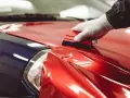 A gloved hand uses a squeegee to apply a shiny red diy car wrap over a blue car fender.