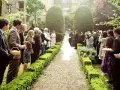 Uroczystość ślubna w ogrodzie. Goście stoją wzdłuż alejki, czekając na rozpoczęcie ceremonii. W tle para młoda i urzędnik.