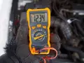 Ręka w rękawicy trzyma pomarańczowy multimetr, który pokazuje 12.34V. Auto nie odpala, akumulator sprawny, problem leży gdzie indziej.